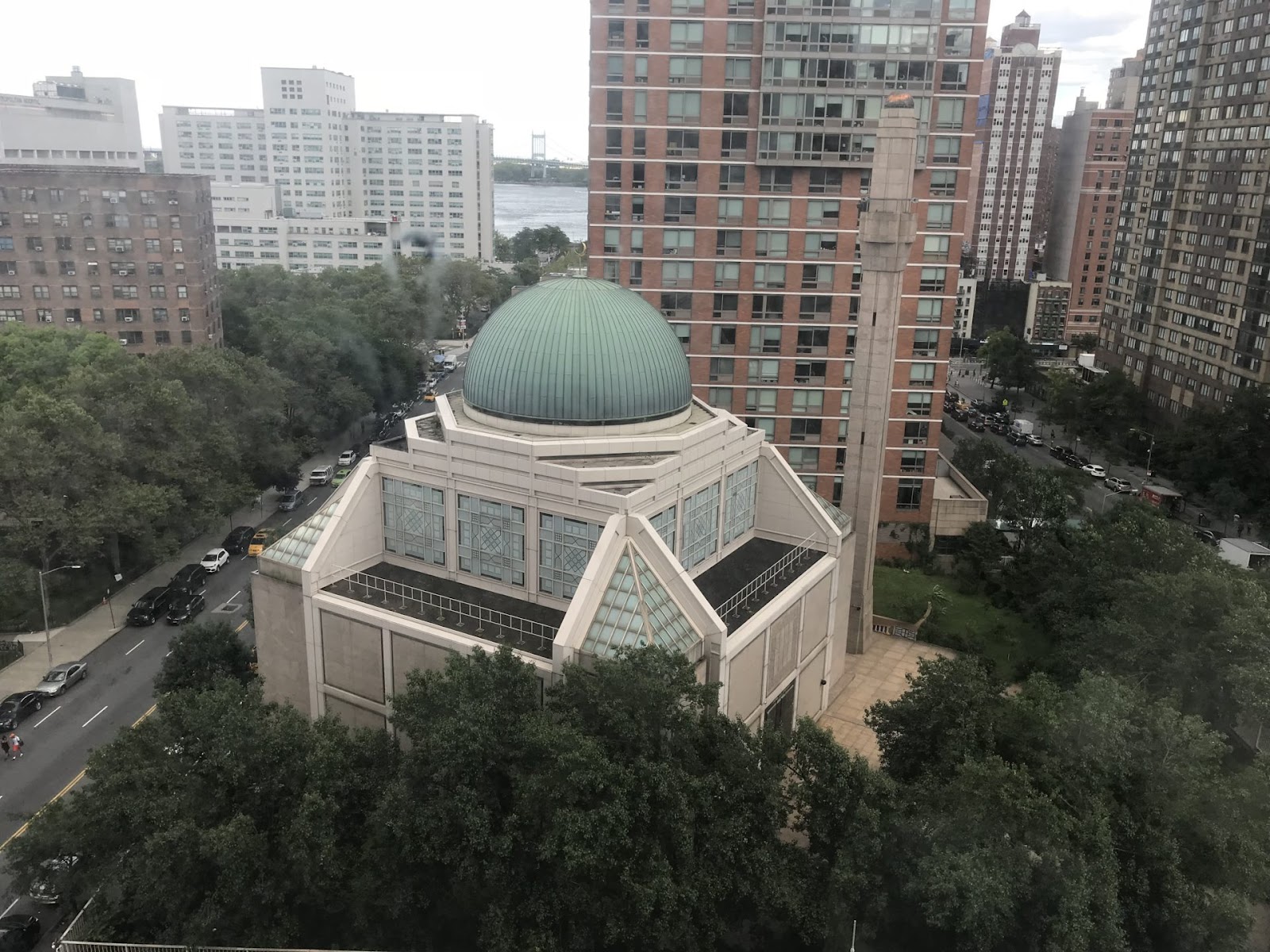 Mint dome that sits on top of a building. this is a famous mosque located in manhattan. lush trees sit at the bottom and behind the mosque you can see city traffic and apartment buildings.